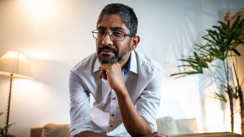 Na imagem, Jeferson Tenório, um homem negro, usando óculos e camiseta branca, posa inclinado sobre uma mesa de madeira, apoiando o queixo na mão direita. Ele tem cabelo curto grisalho, barba e usa óculos. Ao fundo, há um abajur aceso e uma planta, com o ambiente iluminado por luz natural.