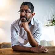 Na imagem, Jeferson Tenório, um homem negro, usando óculos e camiseta branca, posa inclinado sobre uma mesa de madeira, apoiando o queixo na mão direita. Ele tem cabelo curto grisalho, barba e usa óculos. Ao fundo, há um abajur aceso e uma planta, com o ambiente iluminado por luz natural.