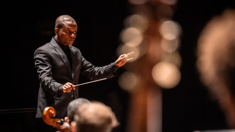 Na imagem, o maestro Anderson Alves, um homem negro, veste um terno preto, segurando a batuta com a mão direita, em um gesto de condução. O fundo é escuro. No primeiro plano, partes desfocadas de instrumentos de corda compõem a cena.