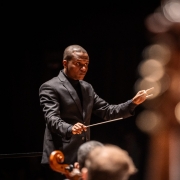 Na imagem, o maestro Anderson Alves, um homem negro, veste um terno preto, segurando a batuta com a mão direita, em um gesto de condução. O fundo é escuro. No primeiro plano, partes desfocadas de instrumentos de corda compõem a cena.