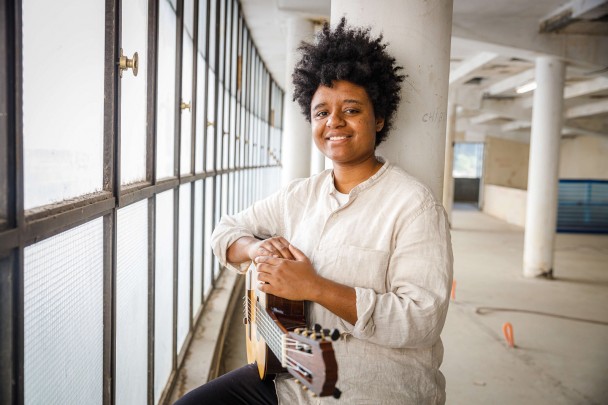 Ao fundo, grandes janelas de vidro divididas por molduras de metal, com colunas brancas e piso claro. Ao centro da imagem, está Gabriele Leite, uma mulher negra, com cabelos crespos, vestindo uma camisa de linho bege, encostada em uma coluna branca, segurando um violão.