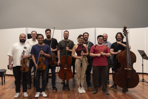 Em um espaço interno com piso de madeira e fundo branco, um grupo de dez músicos posa sorrindo para a foto, com seus instrumentos musicais. Na fileira da frente, quatro homens seguram violinos e violas, ao lado de uma mulher com um oboé e um homem com baquetas de percussão. Na fileira de trás, estão um homem mais velho, sorrindo, e outros quatro músicos, incluindo um flautista e um contrabaixista à direita. Ao fundo, um piano é parcialmente visível.