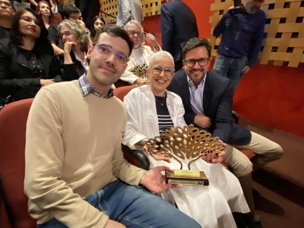 A partir da esquerda, um homem (Benhur), uma mulher (Beatriz) e um homem (Silvio), todos em pé, segurando o troféu do Prêmio Jacarandá