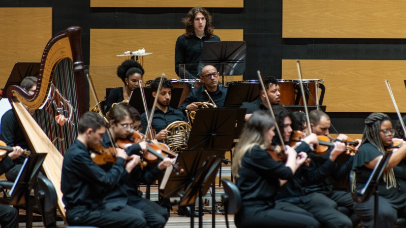 No fundo, painéis de madeira clara com acabamento acústico cobrem a parede, conferindo um ambiente de auditório ou sala de concertos. Ao centro da imagem, músicos da Ospa Jovem estão posicionados em semicírculo, tocando diferentes instrumentos. Em destaque ao fundo, um percussionista de cabelos longos está em pé atrás de um conjunto de tímpanos, enquanto outro músico de percussão está à sua esquerda, parcialmente visível. Mais à frente, há músicos tocando violinos, violoncelos, harpas e instrumentos de sopro, como trompas. Todos vestem trajes formais pretos.