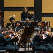 No fundo, painéis de madeira clara com acabamento acústico cobrem a parede, conferindo um ambiente de auditório ou sala de concertos. Ao centro da imagem, músicos da Ospa Jovem estão posicionados em semicírculo, tocando diferentes instrumentos. Em destaque ao fundo, um percussionista de cabelos longos está em pé atrás de um conjunto de tímpanos, enquanto outro músico de percussão está à sua esquerda, parcialmente visível. Mais à frente, há músicos tocando violinos, violoncelos, harpas e instrumentos de sopro, como trompas. Todos vestem trajes formais pretos.