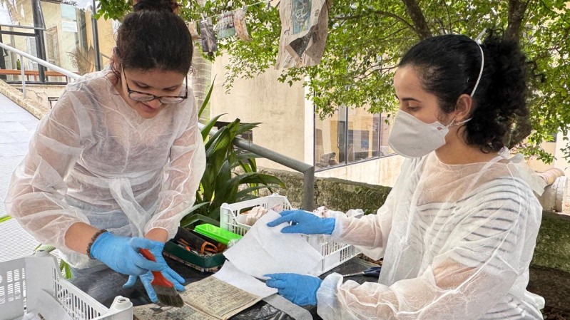 Em um jardim, duas mulheres vestindo equipamentos de proteção individual (EPI) - luvas, avental e máscara, estão separadas por uma mesa coberta com uma lona preta. Sobre a mesa simulam processo de desinfecção de um livro, uma delas segura um pincel enquanto a outra segura um guardanapo.