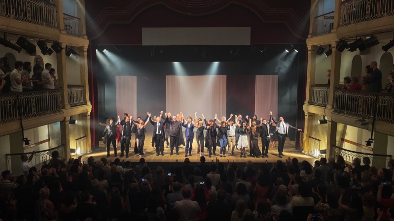 A imagem mostra o concerto de encerramento do Ópera Estúdio em um teatro. No fundo, o palco está iluminado com uma luz suave que realça as paredes vermelhas e as molduras douradas. Ao centro, artistas, incluindo homens e mulheres vestidos formalmente, estão alinhados de mãos dadas e inclinados em reverência ao público, que responde com aplausos. No nível térreo e nas galerias superiores, há uma plateia cheia, com pessoas visivelmente engajadas, algumas em pé, outras tirando fotos ou aplaudindo. A iluminação teatral destaca os artistas e cria uma atmosfera calorosa, enquanto o cenário minimalista ao fundo consiste em cortinas claras que conferem profundidade ao palco.