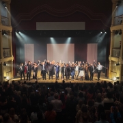 A imagem mostra o concerto de encerramento do Ópera Estúdio em um teatro. No fundo, o palco está iluminado com uma luz suave que realça as paredes vermelhas e as molduras douradas. Ao centro, artistas, incluindo homens e mulheres vestidos formalmente, estão alinhados de mãos dadas e inclinados em reverência ao público, que responde com aplausos. No nível térreo e nas galerias superiores, há uma plateia cheia, com pessoas visivelmente engajadas, algumas em pé, outras tirando fotos ou aplaudindo. A iluminação teatral destaca os artistas e cria uma atmosfera calorosa, enquanto o cenário minimalista ao fundo consiste em cortinas claras que conferem profundidade ao palco.