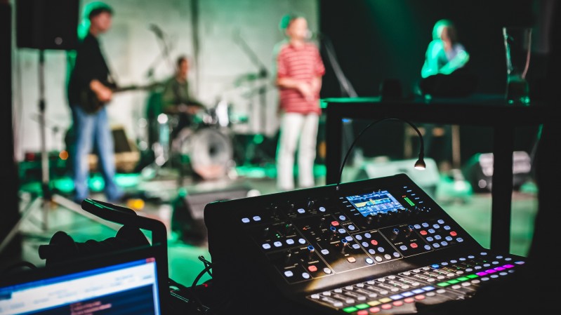 Imagem com fundo desfocado com uma banda em um palco e equipamentos de áudio com botões coloridos no primeiro plano. 