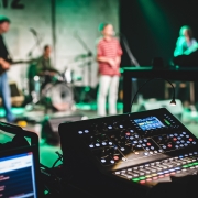 Imagem com fundo desfocado com uma banda em um palco e equipamentos de áudio com botões coloridos no primeiro plano. 