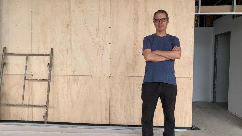 Na foto, o artista Nuno Ramos está em pé e de braços cruzados, em frente a um tapume, olhando para a câmera. Ele é um homem de meia idade, branco, de cabelos grisalhos e usa óculos, camiseta azul, calça e tênis pretos