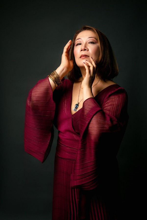 Imagem de Eiko Senda, soprano. Ela está em um retrato artístico com fundo escuro, usando um vestido elegante na cor vinho, de mangas amplas e tecido levemente transparente. Ela exibe uma expressão introspectiva e gesticula delicadamente com as mãos próximas ao rosto. Complementam o visual joias finas, como pulseiras douradas e um colar com um pingente azul.