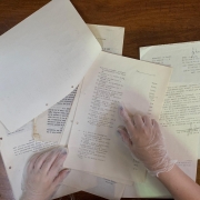 Imagem de uma mesa de madeira com várias folhas de papel datilografadas espalhadas. Algumas folhas possuem texto oficial, com assinaturas e selos, enquanto outras apresentam listas de itens numerados. No canto inferior esquerdo, aparecem duas mãos protegidas por luvas descartáveis, manuseando cuidadosamente os documentos. O ambiente ao fundo é de madeira envernizada, com parte de um móvel curvado visível na parte superior da imagem. No canto superior direito, parte de um pé calçado é parcialmente visível.