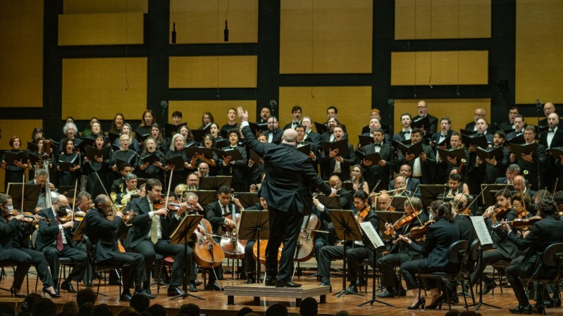 No fundo da imagem, há uma parede de madeira com acabamento em painéis retangulares que compõem o cenário de um auditório. Mais à frente, o Coro da Ospa (Orquestra Sinfônica de Porto Alegre) está posicionado em uma ampla formação, com integrantes segurando partituras e cantando. No centro, em destaque, está o maestro Manfredo Schmiedt, de costas para a câmera, conduzindo a apresentação com gestos expressivos. Na parte frontal, a orquestra está disposta com músicos tocando diversos instrumentos de cordas, como violinos, violas e violoncelos. Os músicos usam trajes formais, e o público, parcialmente visível no canto inferior da imagem, assiste atentamente à performance.