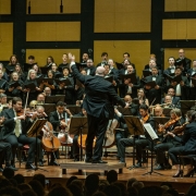 No fundo da imagem, há uma parede de madeira com acabamento em painéis retangulares que compõem o cenário de um auditório. Mais à frente, o Coro da Ospa (Orquestra Sinfônica de Porto Alegre) está posicionado em uma ampla formação, com integrantes segurando partituras e cantando. No centro, em destaque, está o maestro Manfredo Schmiedt, de costas para a câmera, conduzindo a apresentação com gestos expressivos. Na parte frontal, a orquestra está disposta com músicos tocando diversos instrumentos de cordas, como violinos, violas e violoncelos. Os músicos usam trajes formais, e o público, parcialmente visível no canto inferior da imagem, assiste atentamente à performance.