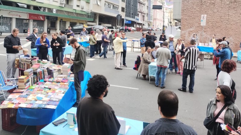 Em uma rua de Porto Alegre, à direita, está a entrada de um prédio de arquitetura moderna. A parte superior direita mostra um cartaz com arte vibrante. Algumas das mesas cobrem uma tela retangular com livros dispostos. Adultos aparecem discutindo e trocando volumes.