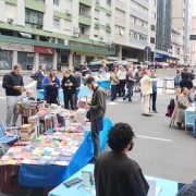 Em uma rua de Porto Alegre, à direita, está a entrada de um prédio de arquitetura moderna. A parte superior direita mostra um cartaz com arte vibrante. Algumas das mesas cobrem uma tela retangular com livros dispostos. Adultos aparecem discutindo e trocando volumes.