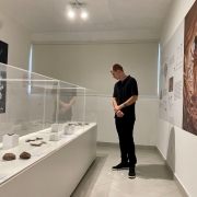 A imagem mostra um homem em uma sala de exposições com diversos paineis fixados nas paredes. Ele observa peças arqueológicas dispostas em expositores no centro da sala. 