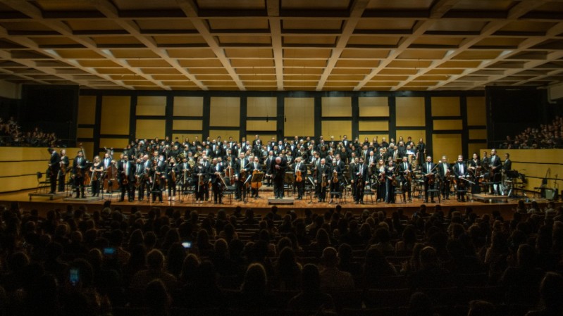 A imagem mostra um concerto da Orquestra Sinfônica, com músicos em pé no palco de uma grande sala de concertos. Os integrantes estão alinhados em diversas fileiras e vestidos formalmente, predominando trajes pretos. O maestro está no centro, à frente, curvando-se em agradecimento ao público. Ao fundo, há um coro posicionado em arquibancadas elevadas. A plateia, em primeiro plano, está parcialmente visível no escuro, com várias pessoas sentadas e assistindo à apresentação. O teto apresenta um design geométrico com painéis retangulares, e as paredes são revestidas de madeira com detalhes modernos.