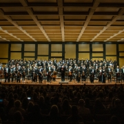 A imagem mostra um concerto da Orquestra Sinfônica, com músicos em pé no palco de uma grande sala de concertos. Os integrantes estão alinhados em diversas fileiras e vestidos formalmente, predominando trajes pretos. O maestro está no centro, à frente, curvando-se em agradecimento ao público. Ao fundo, há um coro posicionado em arquibancadas elevadas. A plateia, em primeiro plano, está parcialmente visível no escuro, com várias pessoas sentadas e assistindo à apresentação. O teto apresenta um design geométrico com painéis retangulares, e as paredes são revestidas de madeira com detalhes modernos.