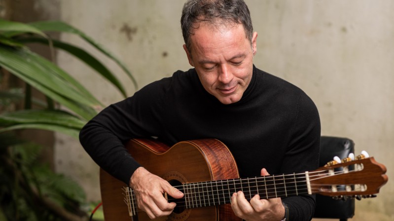 No fundo, um ambiente ao ar livre com folhagens verdes e uma parede com tons de bege e textura rústica. Ao centro, Rodrigo Nassif, homem branco, de cabelos curtos escuros, vestindo um suéter preto, sentado enquanto toca um violão de madeira. Ele está levemente inclinado para frente, com um olhar concentrado e tranquilo, as mãos posicionadas com precisão nas cordas do instrumento.