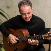 No fundo, um ambiente ao ar livre com folhagens verdes e uma parede com tons de bege e textura rústica. Ao centro, Rodrigo Nassif, homem branco, de cabelos curtos escuros, vestindo um suéter preto, sentado enquanto toca um violão de madeira. Ele está levemente inclinado para frente, com um olhar concentrado e tranquilo, as mãos posicionadas com precisão nas cordas do instrumento.