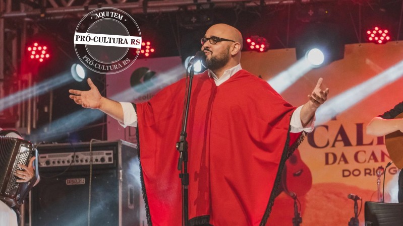 Um homem trajado com roupa típica gaúcha na cor vermelha, de braços abertos, canta no palco da Califórnia da Canção Nativa