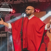 Um homem trajado com roupa típica gaúcha na cor vermelha, de braços abertos, canta no palco da Califórnia da Canção Nativa