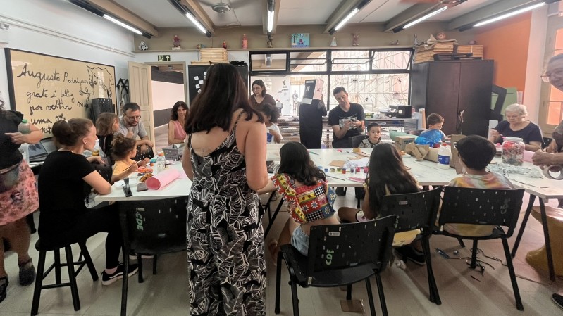 A imagem mostra uma sala iluminada com crianças e adultos reunidos em torno de uma mesa, participando de uma atividade criativa com materiais como tintas, pincéis e rolos de papel. Ao fundo, há estantes com livros, armários e uma parede laranja com grandes janelas. No centro, uma mulher de vestido longo caminha em direção à mesa, enquanto outra, à esquerda, segura um celular. 