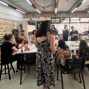 A imagem mostra uma sala iluminada com crianças e adultos reunidos em torno de uma mesa, participando de uma atividade criativa com materiais como tintas, pincéis e rolos de papel. Ao fundo, há estantes com livros, armários e uma parede laranja com grandes janelas. No centro, uma mulher de vestido longo caminha em direção à mesa, enquanto outra, à esquerda, segura um celular. 