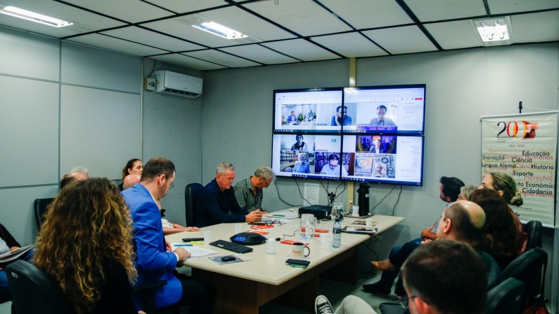 A imagem mostra diversas pessoas em torno de uma mesa em uma sala de reuniões. Em uma das estremidades da mesa, mostra uma tela formada por quatro monitores que mostram imagem de pessoas que participam por videoconferência da reunião. 