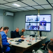 A imagem mostra diversas pessoas em torno de uma mesa em uma sala de reuniões. Em uma das estremidades da mesa, mostra uma tela formada por quatro monitores que mostram imagem de pessoas que participam por videoconferência da reunião. 