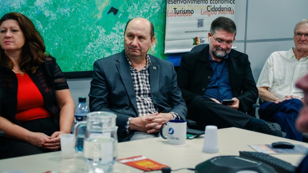 A imagem mostra o deputado Elton Weber ao centro da imagem em uma sala de reuniões, com um homem a sua esquerda. Ao fundo, a imagem mostra a parte inferior de um banner do Bicentenário em que aparecem as palavras turismo, educação, cidadania e lingua alemã.