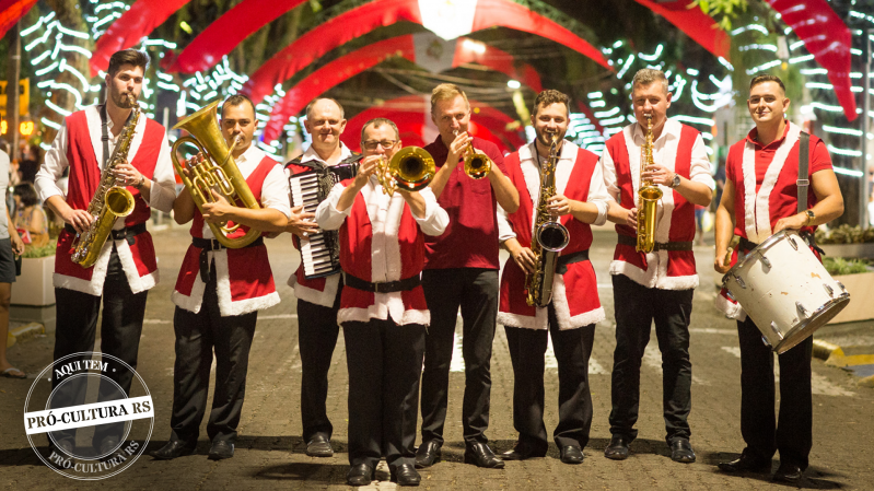 Oito homens com trajes natalinos estão em pé, lado a lado, tocando instrumentos de música típica alemã, em uma avenida enfeitada com decoração de Natal