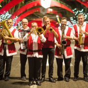 Oito homens com trajes natalinos estão em pé, lado a lado, tocando instrumentos de música típica alemã, em uma avenida enfeitada com decoração de Natal