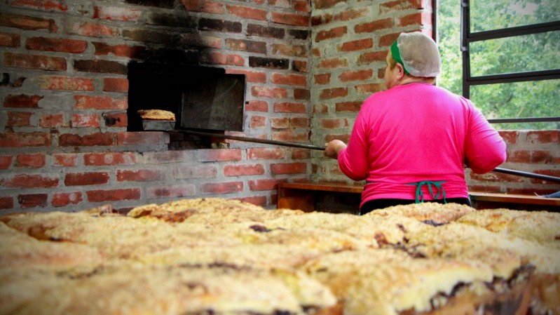 Ao fundo, uma cuqueira de costas coloca uma cuca para assar em um forno de pedra. À frente, há várias cucas, uma ao lado da outra