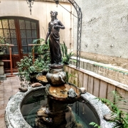 Jardim de inverno da Biblioteca Pública do Estado (BPE), com uma fonte central feita de pedra, adornada por uma estátua de mulher em posição elegante no topo. A fonte é rodeada por pedras decorativas e plantas em vasos. Ao fundo, portas de madeira com vidro fosco, divididas em quadrados, se abrem para salas internas.