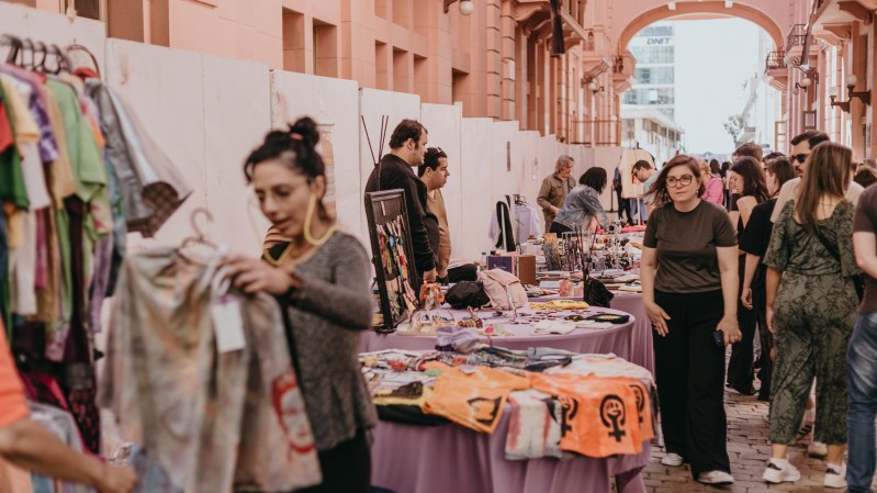 A imagem mostra uma feira acontecendo na Travessa Rua dos Cataventos, na CCMQ, com diversas mesas, nas quais há produtos expostos. Atrás da mesa, estão os expositores e, à frente, os visitantes