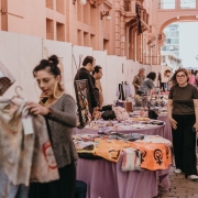 A imagem mostra uma feira acontecendo na Travessa Rua dos Cataventos, na CCMQ, com diversas mesas, nas quais há produtos expostos. Atrás da mesa, estão os expositores e, à frente, os visitantes