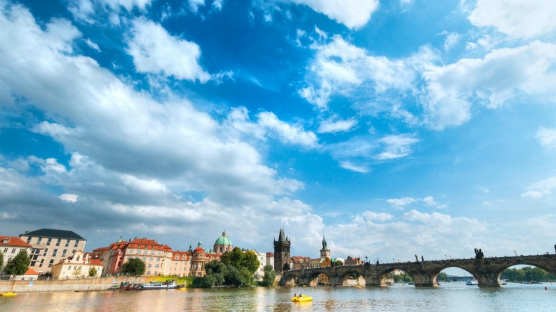 Fotografia de Praga, na República Tcheca. À frente aparece um rio e, ao fundo, uma ponte e prédios históricos