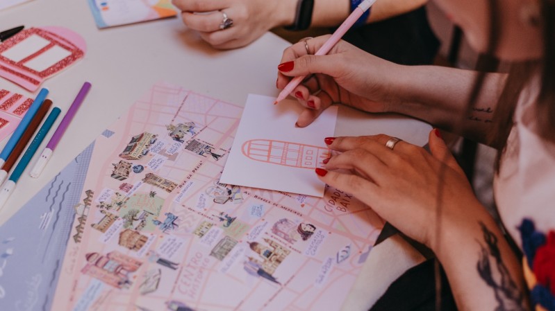 A imagem mostra as mãos de uma pessoa desenhando sobre uma mesa.