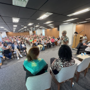 Em um palco, quatro pessoas estão sentadas e uma delas está em pé, ao microfone. É a secretária Beatriz Araujo. Ao fundo, o público assiste