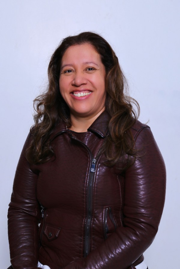 Uma mulher indígena sorri olhando para a câmera. Ela veste casaco cor de vinho