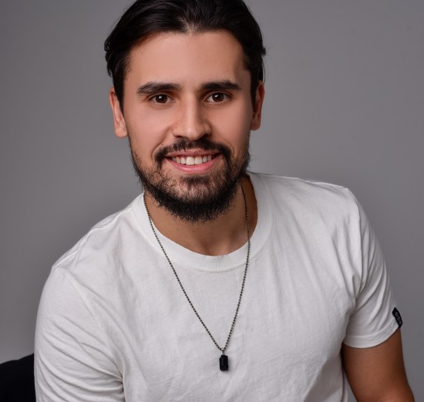 Um homem branco, de barba, sorri olhando para a câmera. Ele veste uma camiseta branca