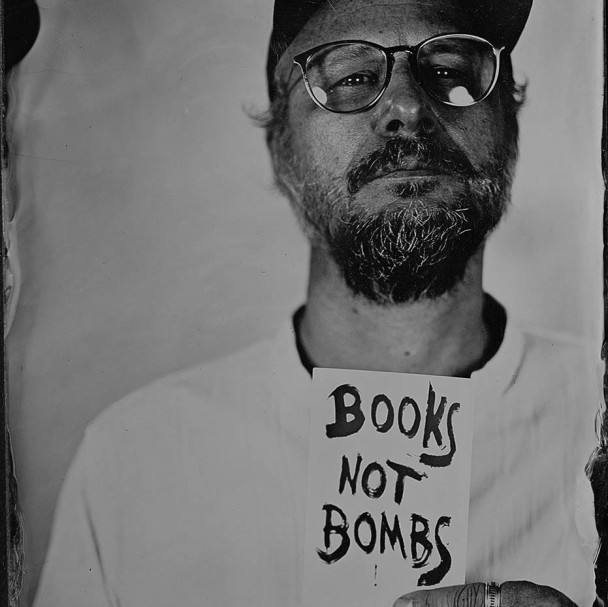 Um homem branco, de óculos, barba e camiseta branca, segura um papel com a frase "Books not bombs"