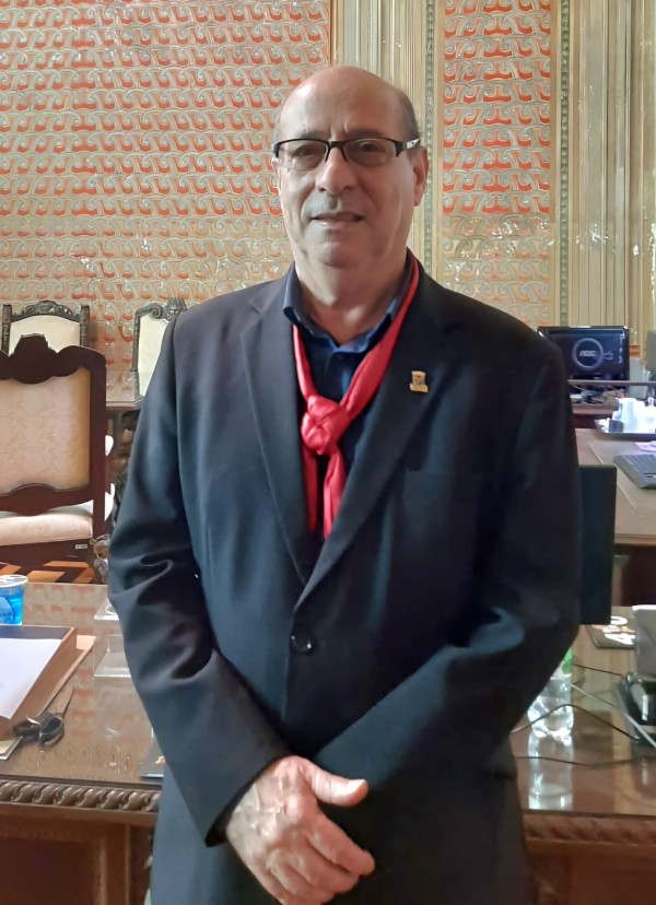 Um homem branco, de óculos, está em pé, de terno preto e lenço vermelho no pescoço