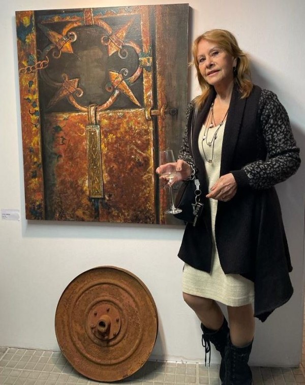 Uma mulher loira está em pé, segurando uma taça, enquanto olha para a câmera e sorri. Ela está ao lado de um quadro