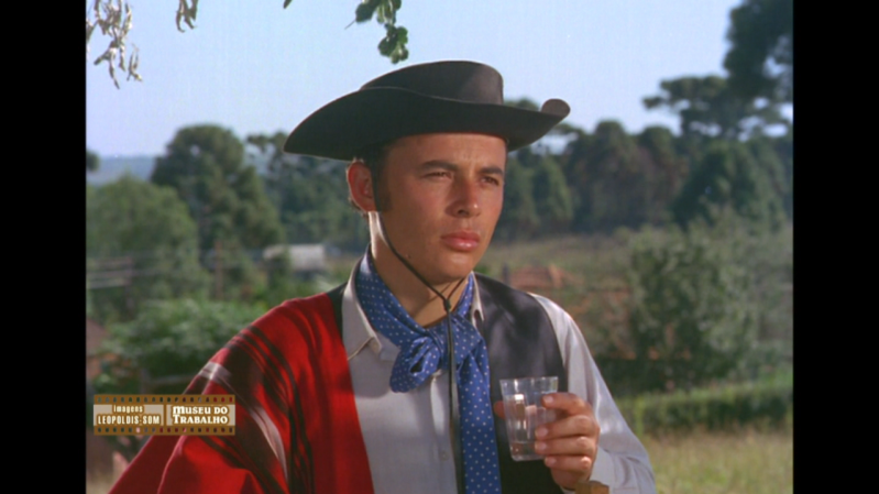 Cena do filme "Pára, Pedro!", com o cantor e ator José Mendes caracterizado como gaúcho. Ele veste um chapéu preto, lenço azul de bolinhas, camisa branca, colete preto e um poncho vermelho e preto sobre os ombros. Segura um copo de vidro transparente com uma bebida, enquanto olha para o lado com expressão séria. O fundo exibe uma paisagem rural com árvores e céu azul.