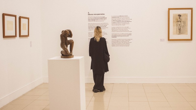 A imagem mostra uma pessoa vestida de preto, de costas, observando um texto informativo em uma parede de galeria de arte. O texto na parede tem o título "Obras recuperadas" e descreve informações sobre a recuperação de obras artísticas. À esquerda da pessoa, há uma escultura de bronze representando uma figura humana musculosa, sentada com uma das pernas dobradas e a cabeça apoiada em um dos braços. A escultura está sobre um pedestal branco. Na parede, ao redor, há várias obras emolduradas, incluindo dois desenhos pequenos à esquerda e um desenho maior de uma figura feminina nua à direita. 