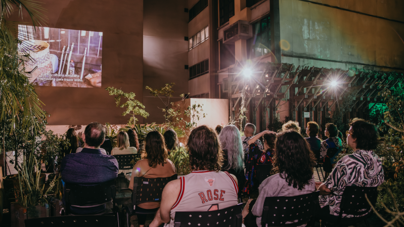 A imagem retrata uma sessão de cinema ao ar livre em um espaço urbano durante a noite. No fundo, há prédios altos com fachadas iluminadas por luzes artificiais, além de uma estrutura de madeira com plantas trepadeiras e iluminação destacada. Algumas plantas e arbustos em vasos estão espalhados pelo local, trazendo um toque de natureza ao ambiente. No centro da imagem, uma projeção é exibida em uma parede clara, mostrando uma cena com utensílios de cozinha e legendas em português. As legendas dizem: "Eu vou olhar a armadilha e aproveito para trazer lenha."
Em primeiro plano, várias pessoas estão sentadas em cadeiras pretas de plástico, voltadas para a projeção.